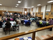 A large multipurpose room filled with exercise equipment in the background. In the foreground, students and adults sit at circular tables covered in blue, pink, and green cloths, focused on writing or crafting.